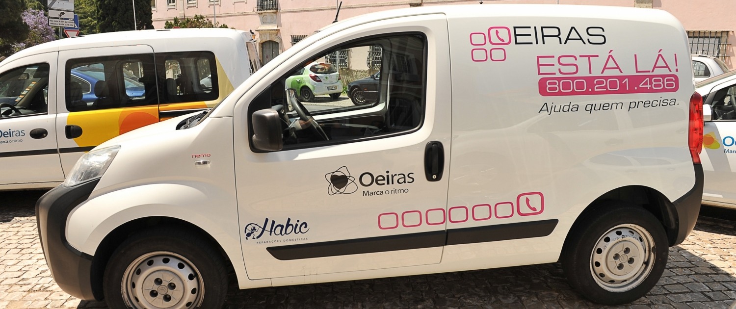 Vista lateral de uma carrinha branca com vários logótipos, entre os quais "Habic" e "Oeiras"